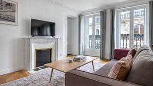 Una sala de estar con chimenea, un televisor de pantalla plana montado encima, un sofá, una mesa de centro con libros y ventanas amplias con vista a un edificio.