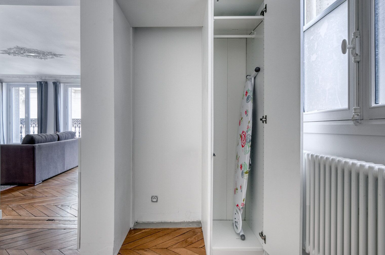 Une pièce dotée d’un parquet en bois disposé en chevrons, de murs blancs, d’un radiateur et d’une fenêtre donnant sur un balcon.