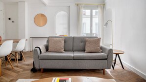 Ein modernes Wohnzimmer mit einem grauen Sofa, weißen Sesseln, einem Holzesstisch und einem runden Spiegel an der Wand.