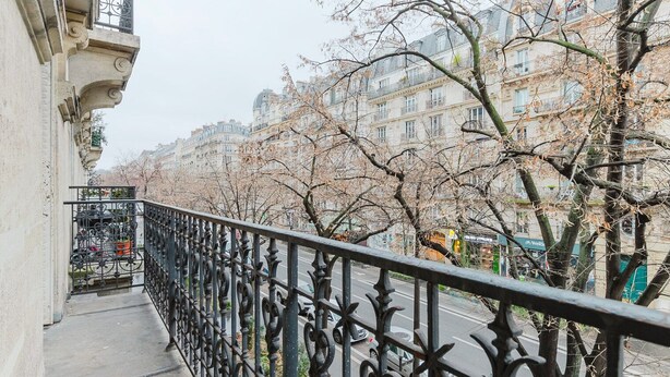 Ein Balkon mit aufwendiger schwarzer Metallgliederung, der auf eine von Bäumen und Gebäuden gesäumte Straße blickt.