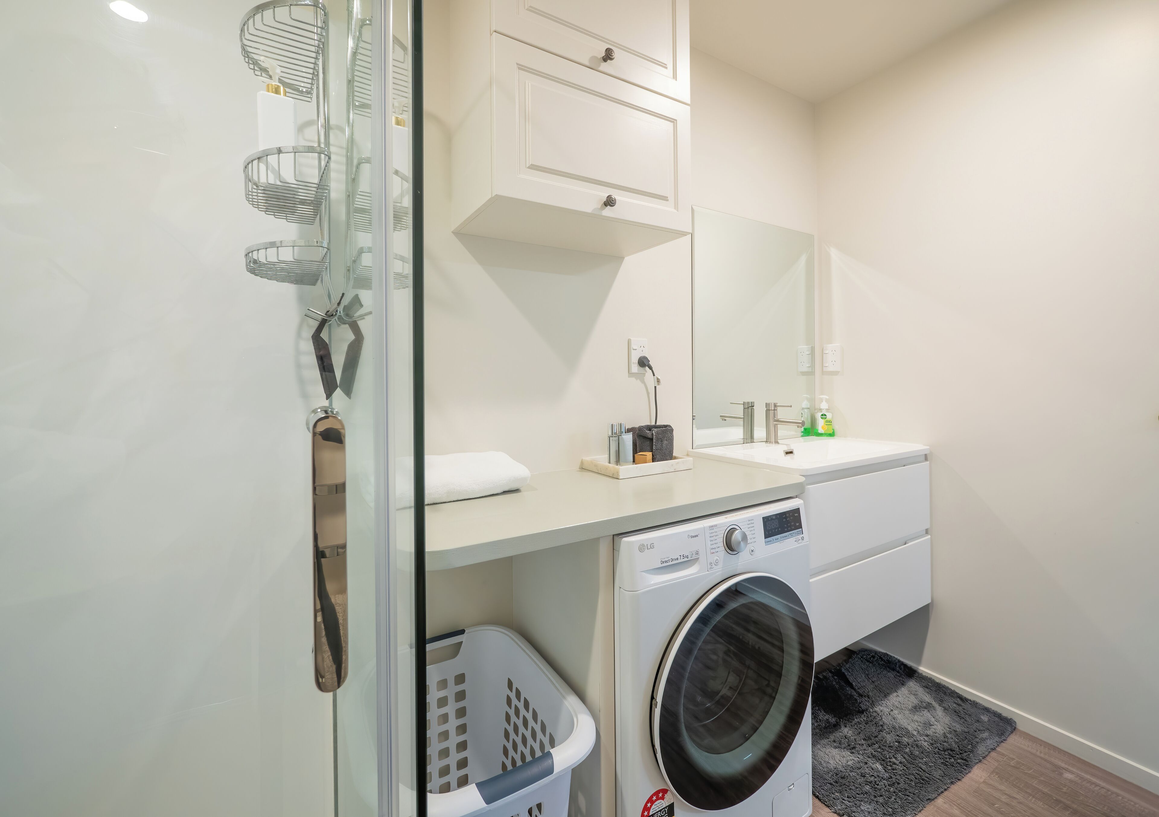 A compact laundry area with a washing machine, a drying rack, a white laundry basket, a mirror, and a small cabinet.