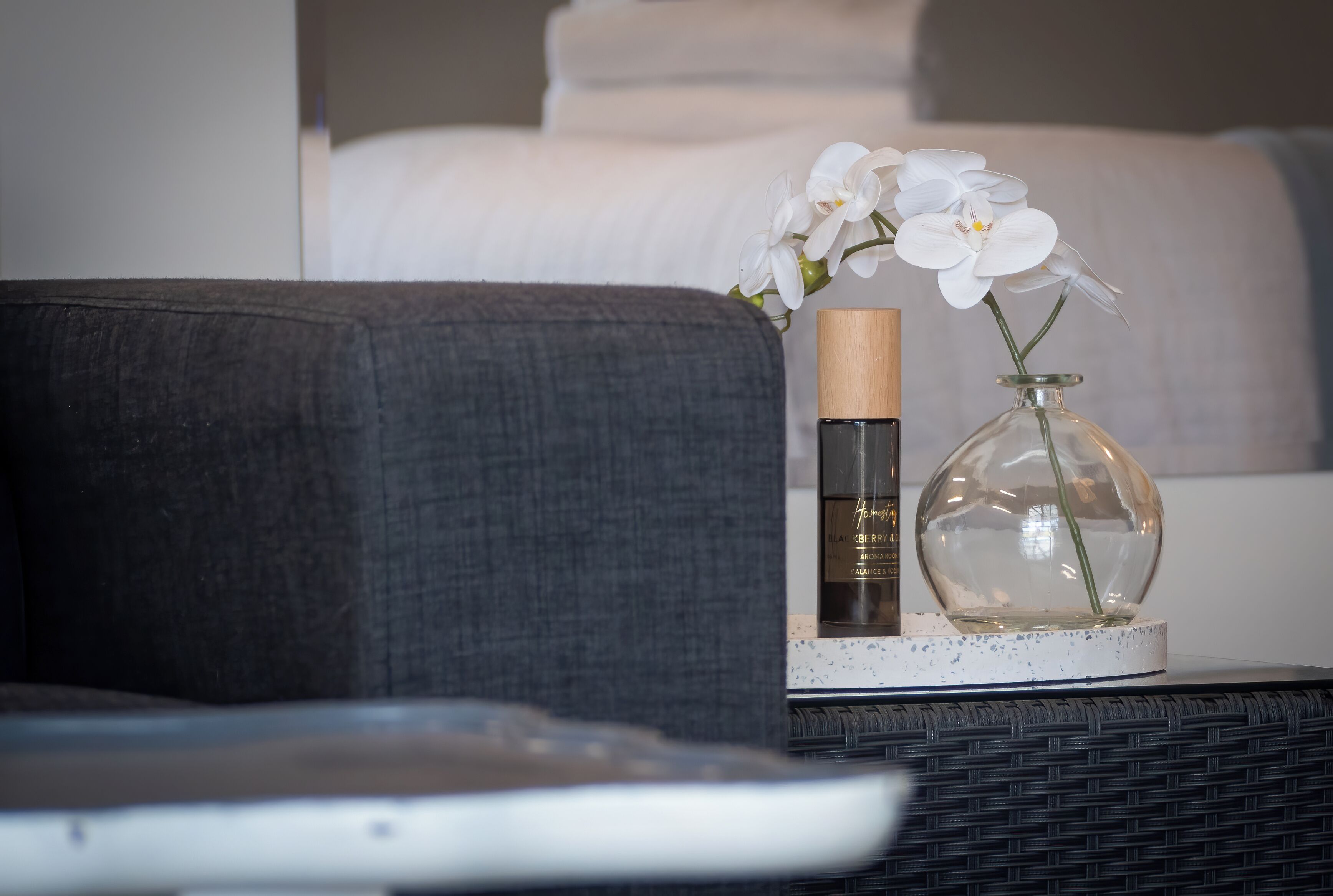 A bedside table with a bottle, a glass vase with a plant, and a grey sofa in the background.