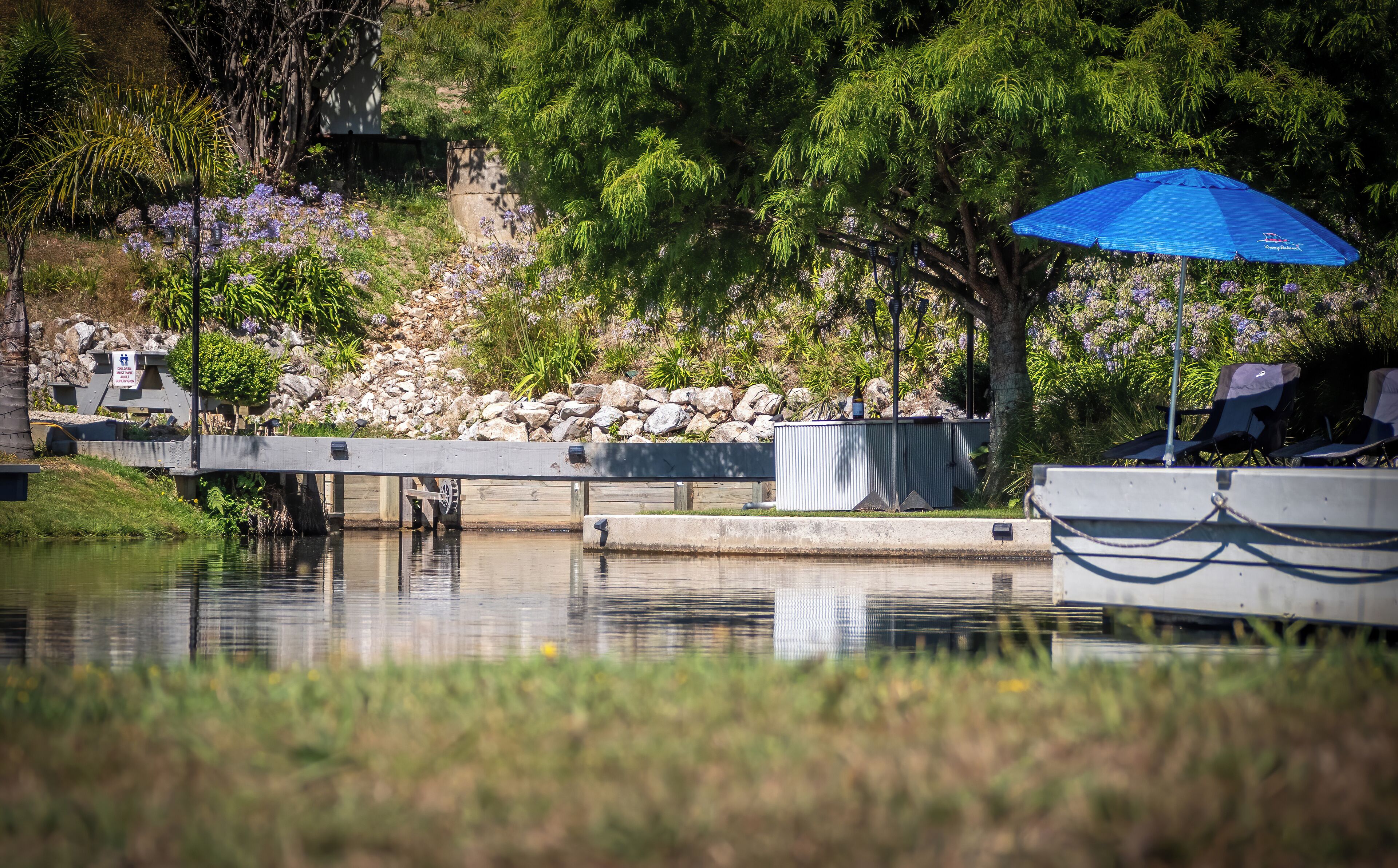 A tranquil riverside setting with a bridge, a concrete barrier, and a blue umbrella providing shade.