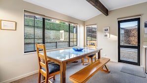 A dining area with a glass-top table, wooden chairs, and a bench. There are large windows with a view of a garden and a door leading outside. - Criffel Peak Cottage, quiet location, only 10 min walk into Wanaka town centre (Wānaka)