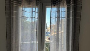 A view through a window with sheer curtains, revealing a balcony and a street scene with buildings and cars. - Nissaki Rooftop Oceanview two bedroom apartment with WiFi and air conditioning (Nisaki)