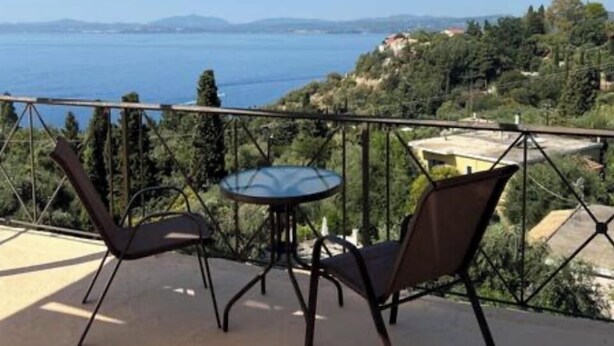 A balcony with two chairs and a table, overlooking a scenic view of the sea and hills. - Nissaki Rooftop Oceanview two bedroom apartment with WiFi and air conditioning (Nisaki)