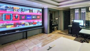 A modern room with a large flat-screen TV displaying various streaming services. There is a desk with a computer and a chair. The room has a tiled floor and a brick accent wall. - Pyeongtaek Station Gallery Hotel (Pyeongtaek)