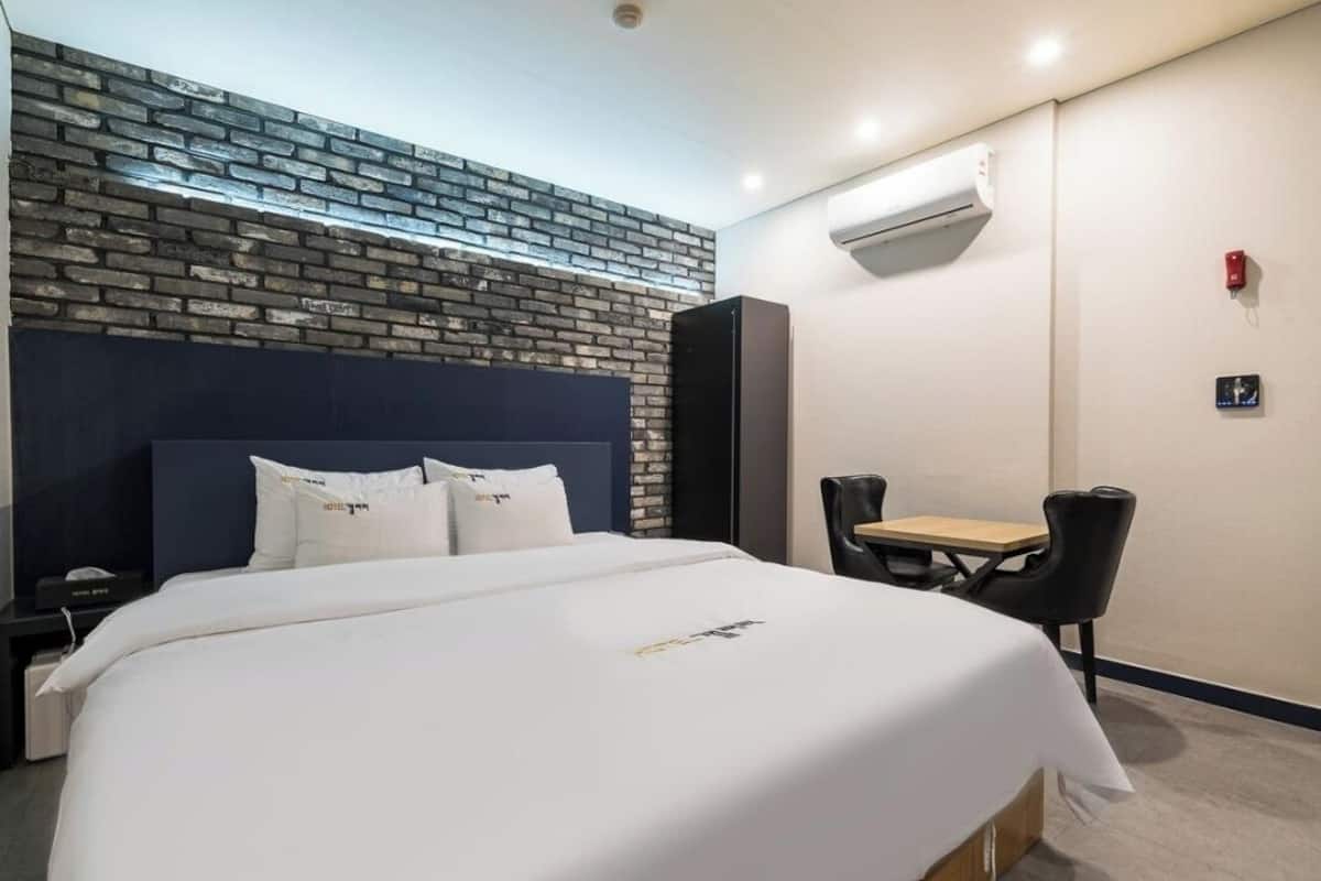Habitación de hotel con cabecera de ladrillo, cama con ropa blanca, una mesita con dos sillas y un equipo de aire acondicionado en la pared.