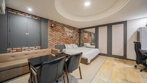 A modern hotel room with a brick accent wall, a large bed, a sofa, a dining table with chairs, and a desk with a chair. - Pyeongtaek Station Gallery Hotel (Pyeongtaek)