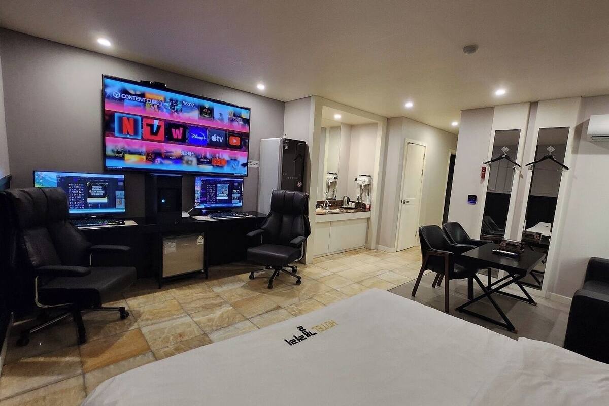 Habitación de hotel con una cama grande, un escritorio con dos computadoras, un televisor mostrando servicios de streaming y un armario con perchas.