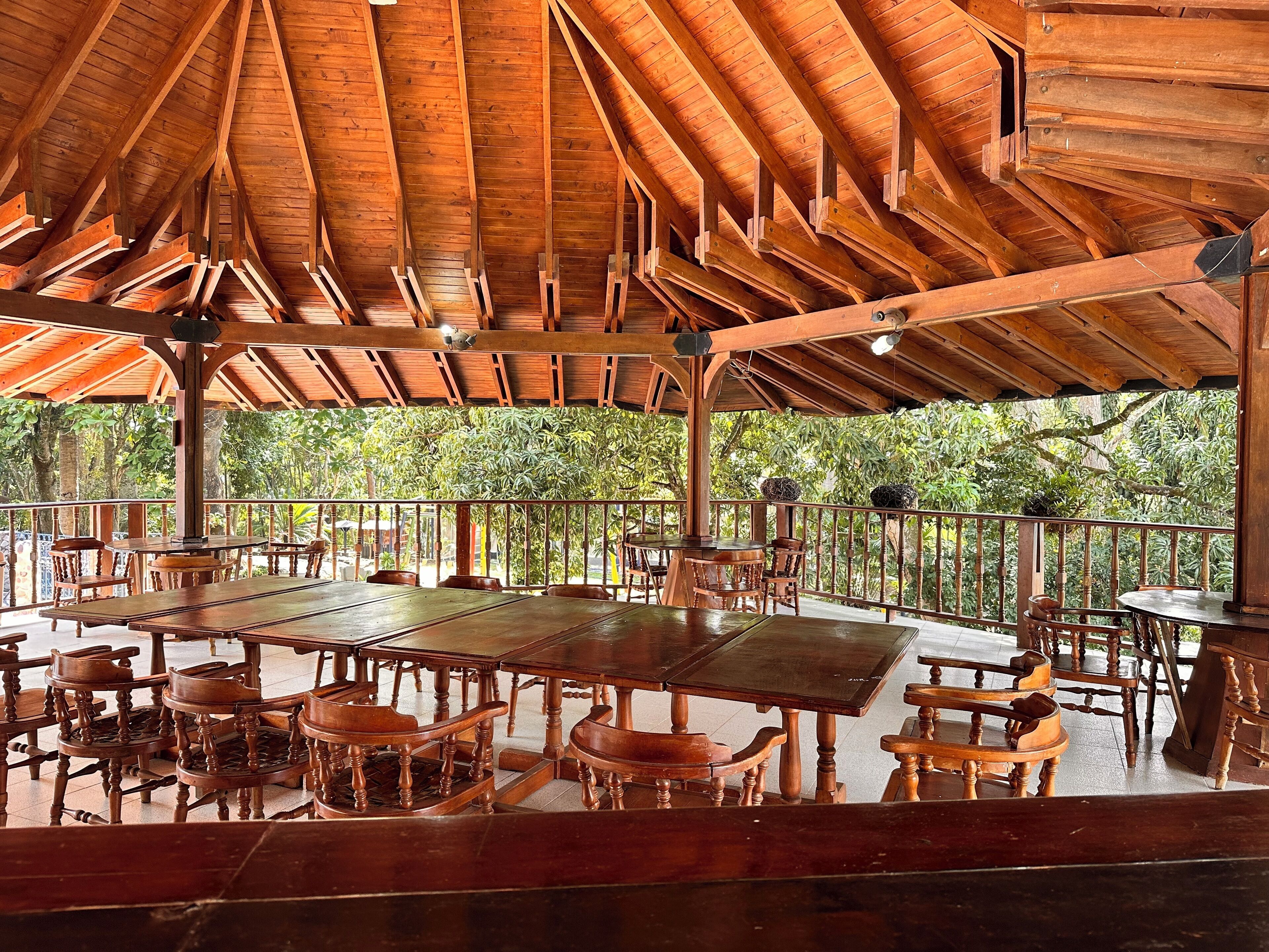 Un espacio de comedor al aire libre cubierto, con mesas y sillas de madera, rodeado por una barandilla y abundante vegetación.