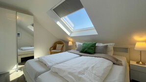 Ein Schlafzimmer mit Dachfenster, einem Bett mit weißer Bettwäsche, einem Sessel und einem Nachttisch mit Lampe.