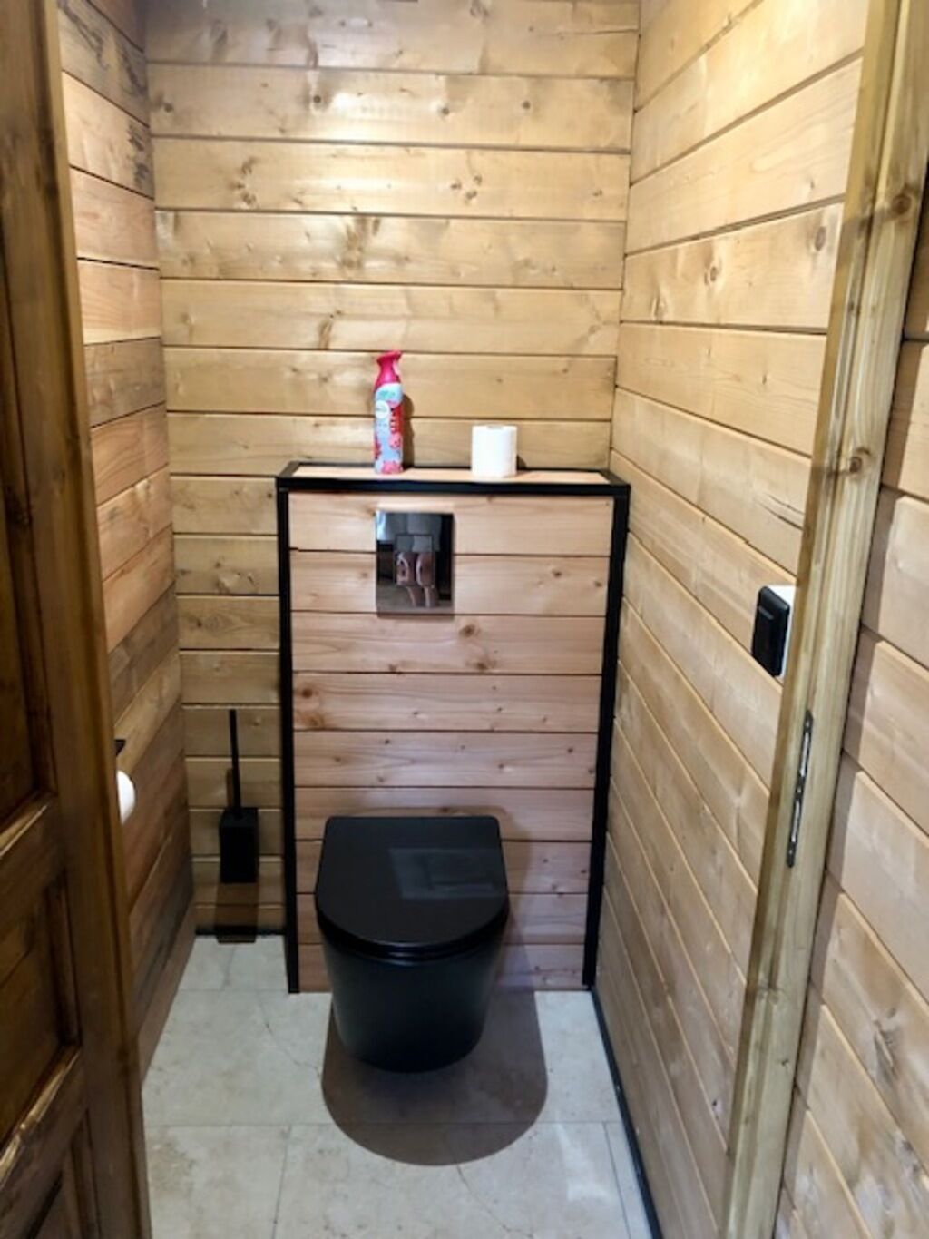 A small, wooden-paneled bathroom with a black toilet, a white toilet paper holder, and a cleaning spray bottle on a shelf.