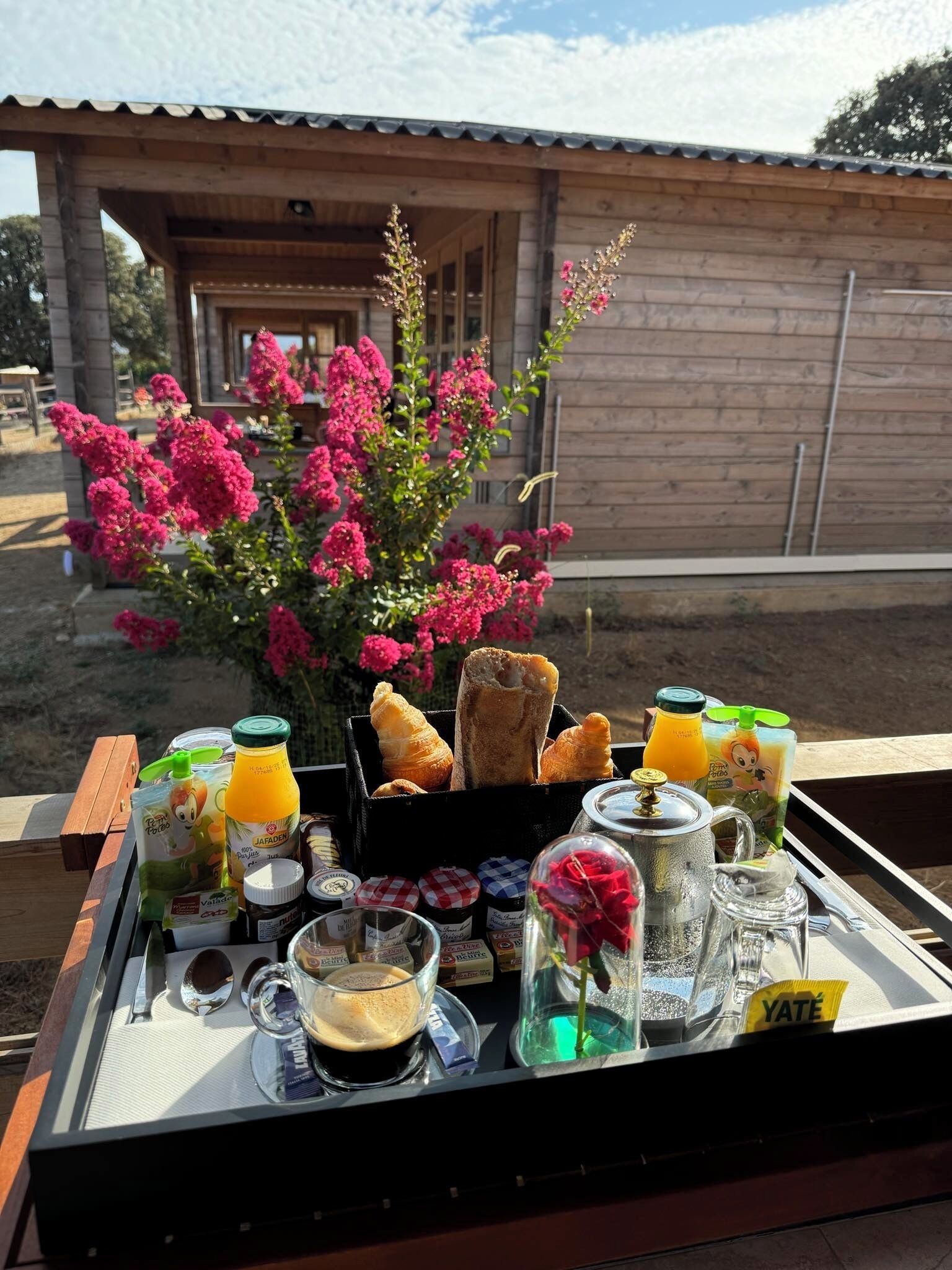 A breakfast tray with croissants, juice, and coffee on a wooden deck with a view of a rustic building.