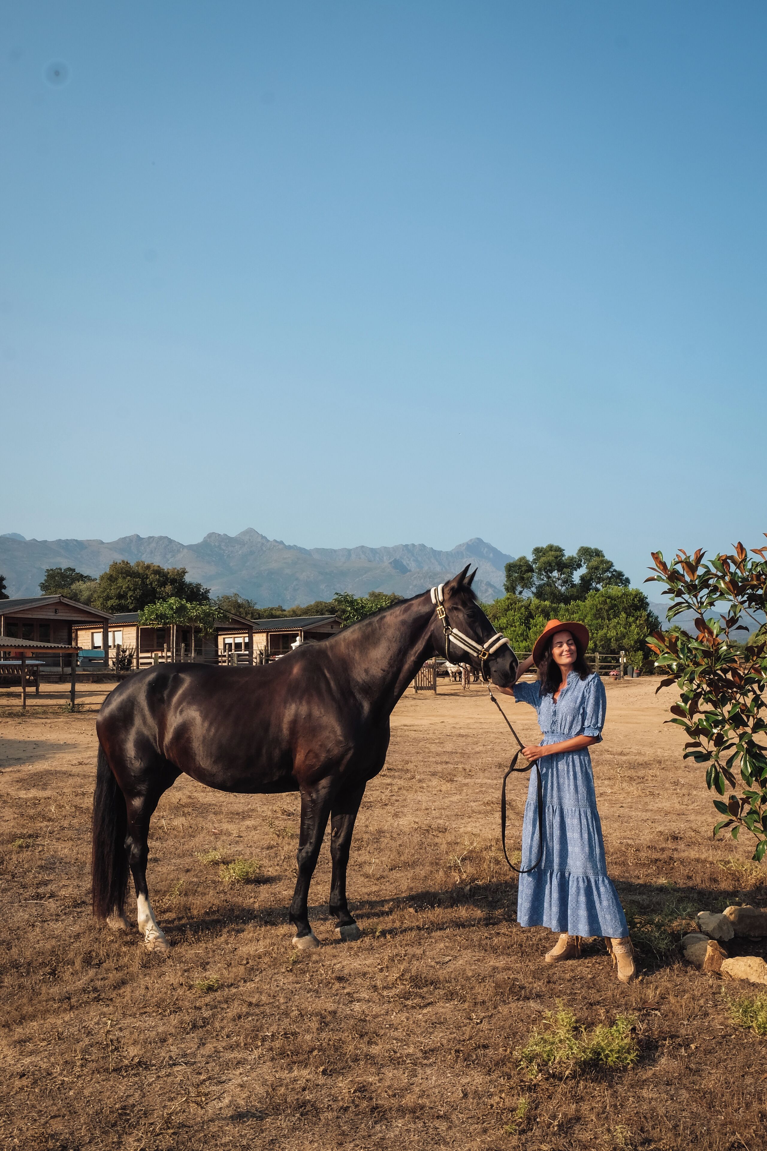 A person in a blue dress standing next to a horse in an outdoor area with buildings and mountains in the background.
