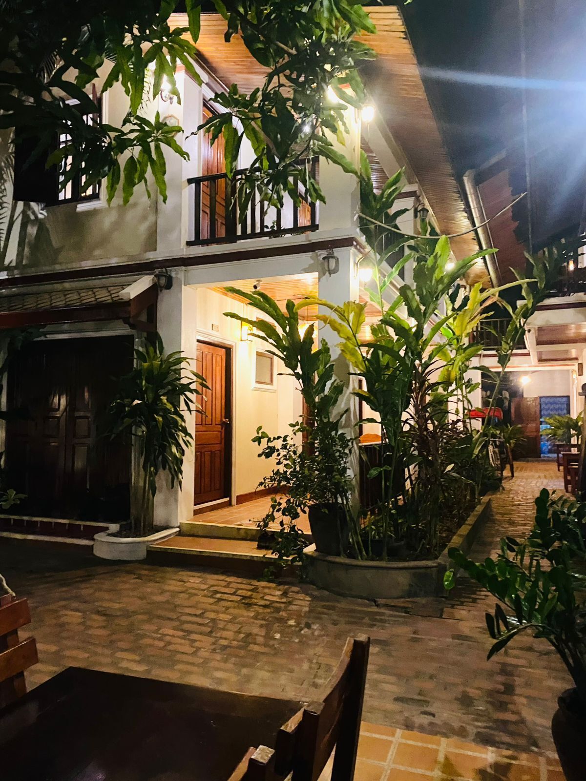 Un patio con mesa y sillas de madera, rodeado de plantas en macetas y un edificio con balcón.