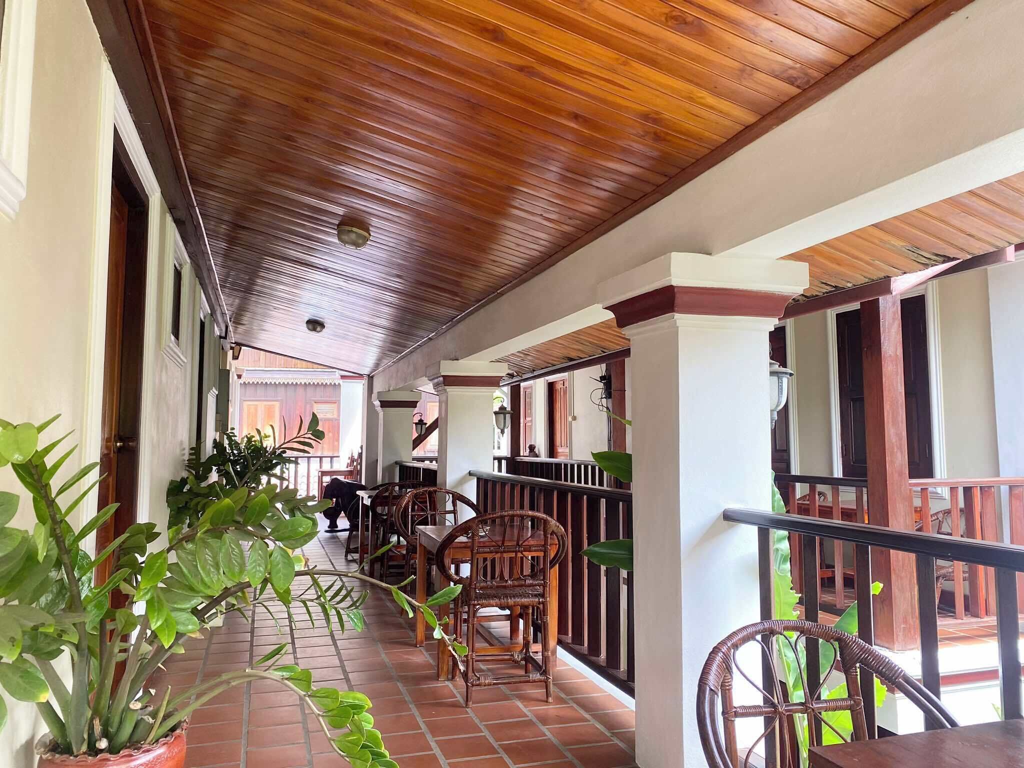 A covered corridor with wooden ceiling and tiled floor, featuring wicker chairs and potted plants.