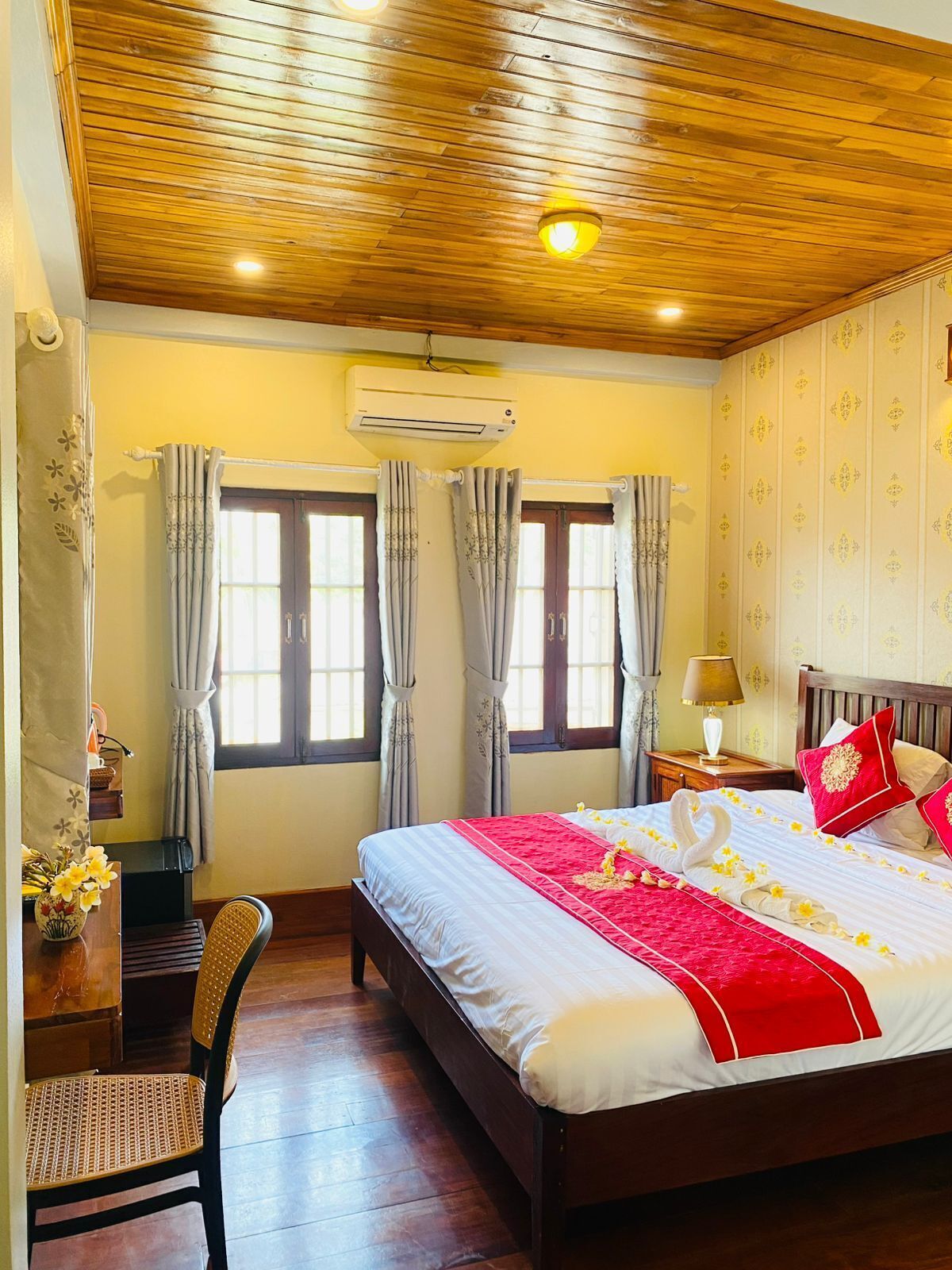 A hotel room with a wooden ceiling, two windows with curtains, a bed with a red and white bedspread, a wooden chair, and a desk with a lamp.