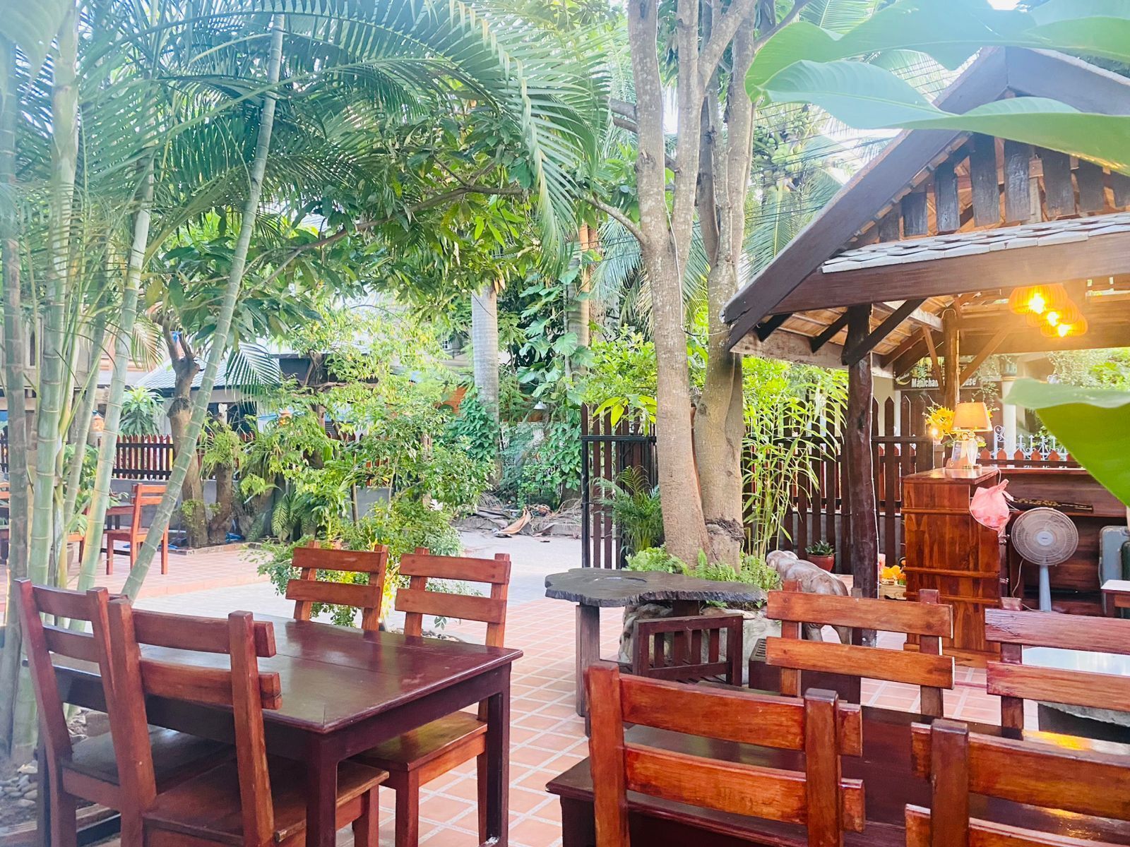 A wooden outdoor seating area with tables and chairs, surrounded by lush greenery and trees.