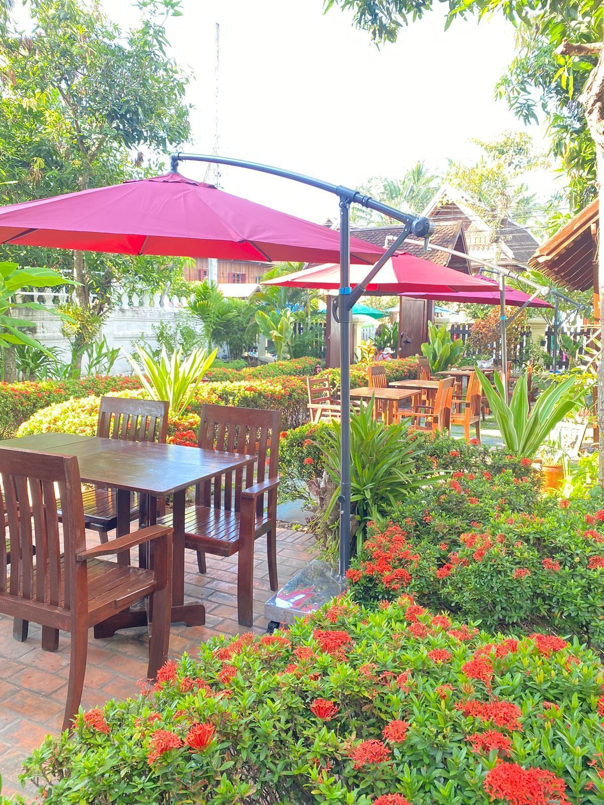 An outdoor seating area with wooden tables and chairs, red umbrellas, and lush greenery.