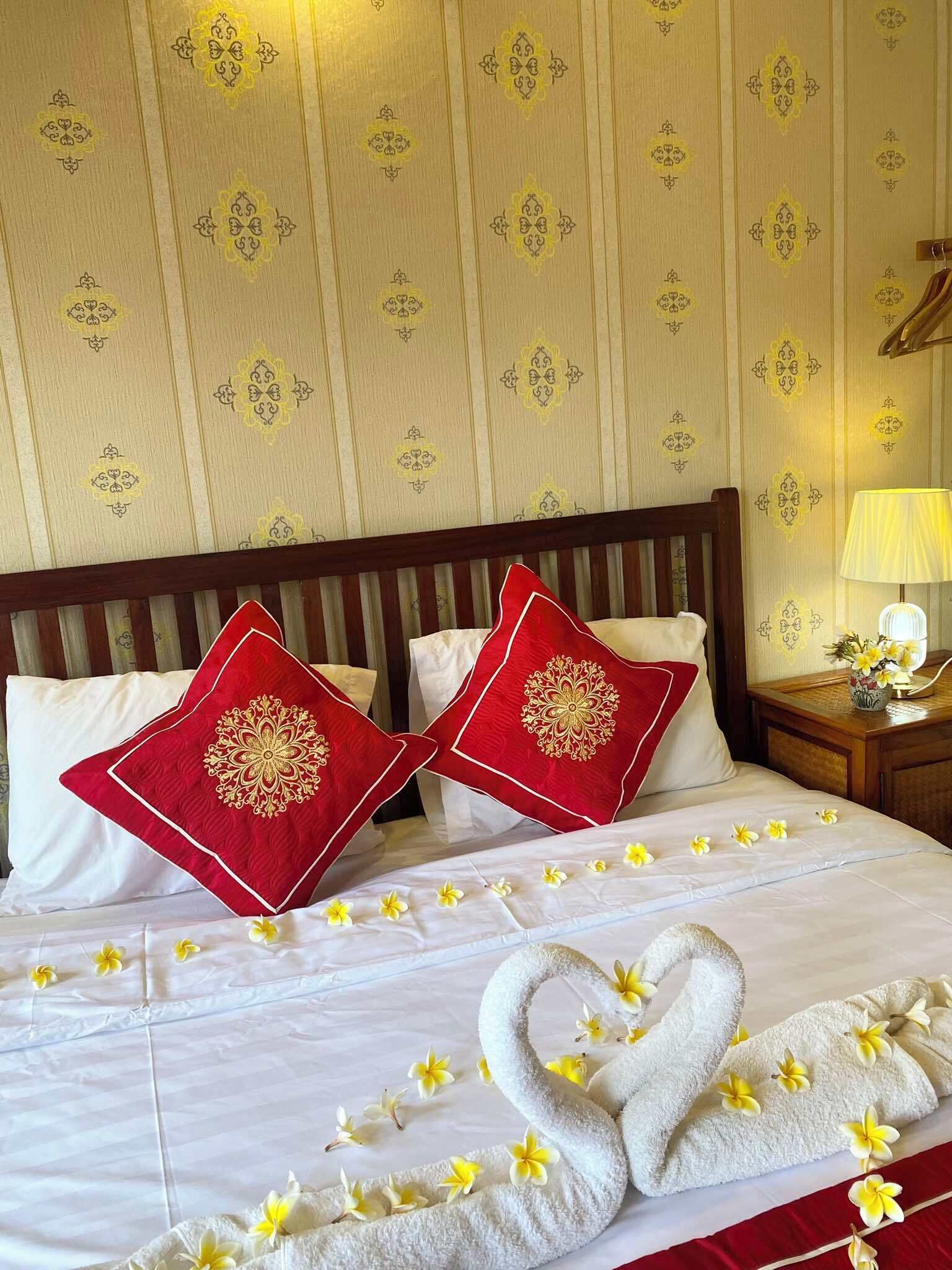 Una habitación de hotel con una cama, dos almohadas rojas con bordado dorado, un cabecero de madera, una mesita de noche con una lámpara y un arreglo floral.