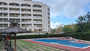 Une piscine bordée de rouge, entourée d’une zone pavée parsemée de quelques touffes d’herbe, d’un parasol en chaume et d’une bouée de sauvetage.