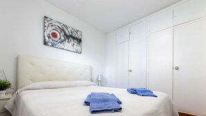Une chambre à coucher avec un lit blanc, une tête de lit, une œuvre d’art accrochée au mur, une table de chevet avec une lampe et une armoire.
