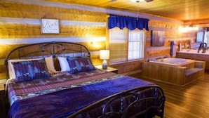 Ein Schlafzimmer in einer Holzhütte mit einem Metallbett, einem großen Fenster mit Jalousien, einer Holzwanne und einem kleinen Badezimmer mit Spiegel und Waschbecken.