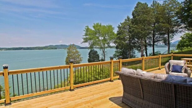 Eine Holzterrasse mit Geländer, mit Blick auf einen See, Bäume und Hügel in der Ferne.