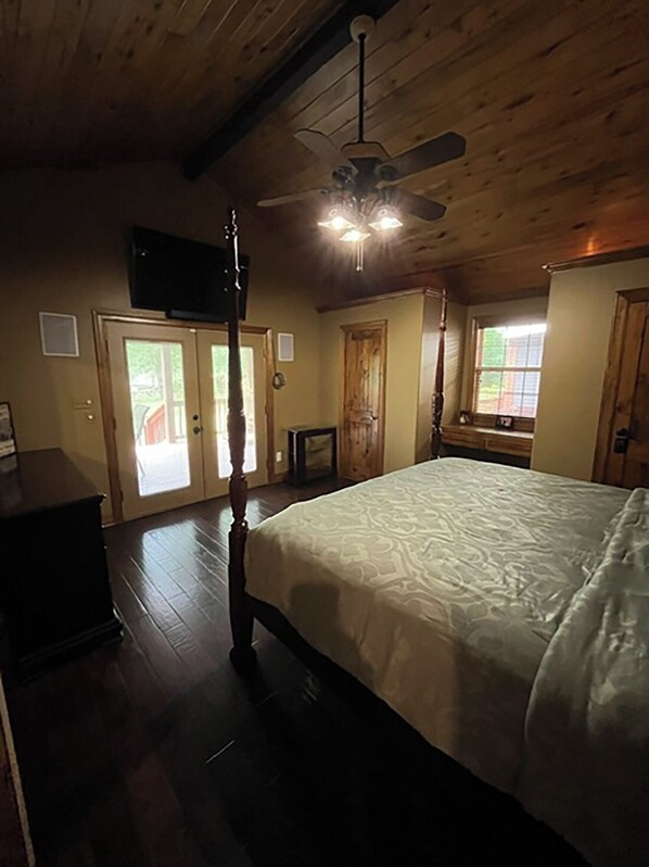 A bedroom with a four-poster bed, wooden floors, a ceiling fan, and a television on the wall.
