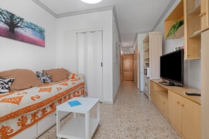 A compact living space with a sofa, a television, a small table, and a hallway leading to a kitchen area.