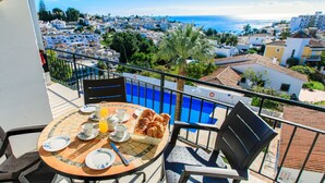 A balcony with a table set for breakfast, overlooking a coastal town and a blue pool. - Florida by Interhome (Nerja)
