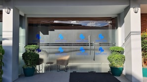 A modern hotel entrance with a glass door featuring the logo 'Rudhil's Place Hotel & Dormitory'. There are two potted plants on either side of the door, and a small table with a chair in front. - Rudhil's Place Hotel (Cebu City)