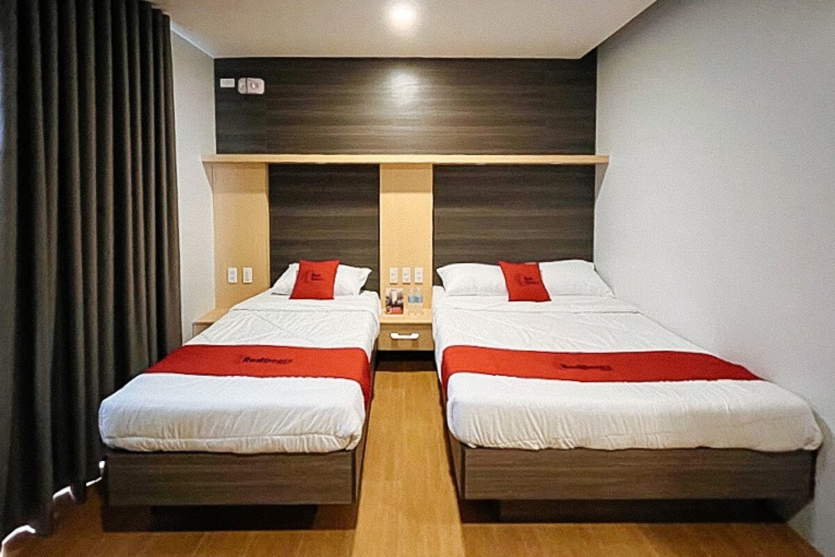 A hotel room with two beds, a wooden headboard, a nightstand, and a red accent pillow.