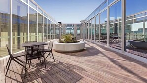 Una terraza moderna al aire libre con paredes de vidrio, piso de madera y un macetero central con follaje.