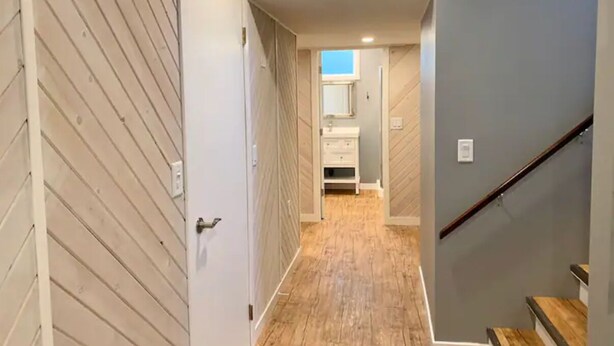 A hallway with wooden flooring, a white door, a staircase with a wooden handrail, and a gray wall. - The Whispering Cedar (Valemount)