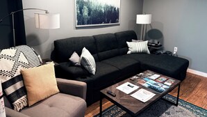A modern living room with a sectional sofa, a coffee table with magazines, and a large forest-themed artwork on the wall. - The Whispering Cedar (Valemount)