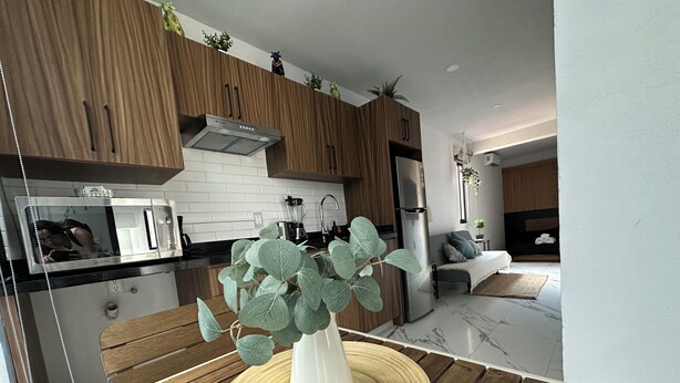 Een moderne keuken met houten kastjes, een inbouwoven en een eethoek met een ronde houten tafel en een witte vaas met groene bladeren.