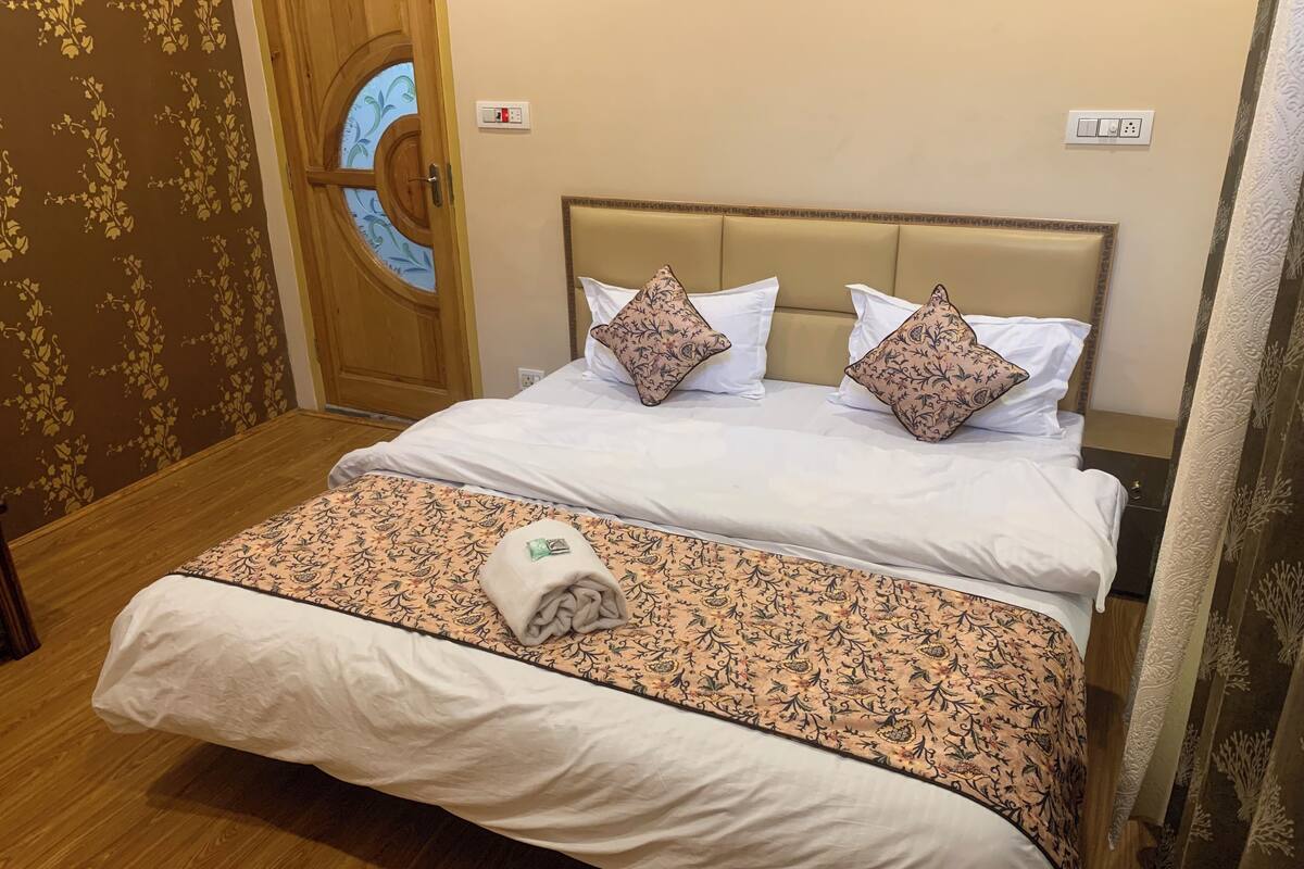 A hotel room with a bed, two pillows, a blanket, a towel, a wooden door, and a wall with a power outlet.