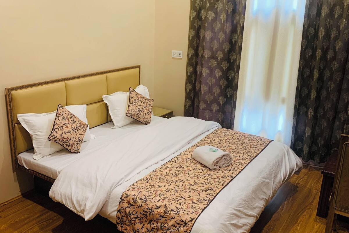 A neatly made bed with a patterned bedspread and pillows, a wooden floor, and a window with curtains.
