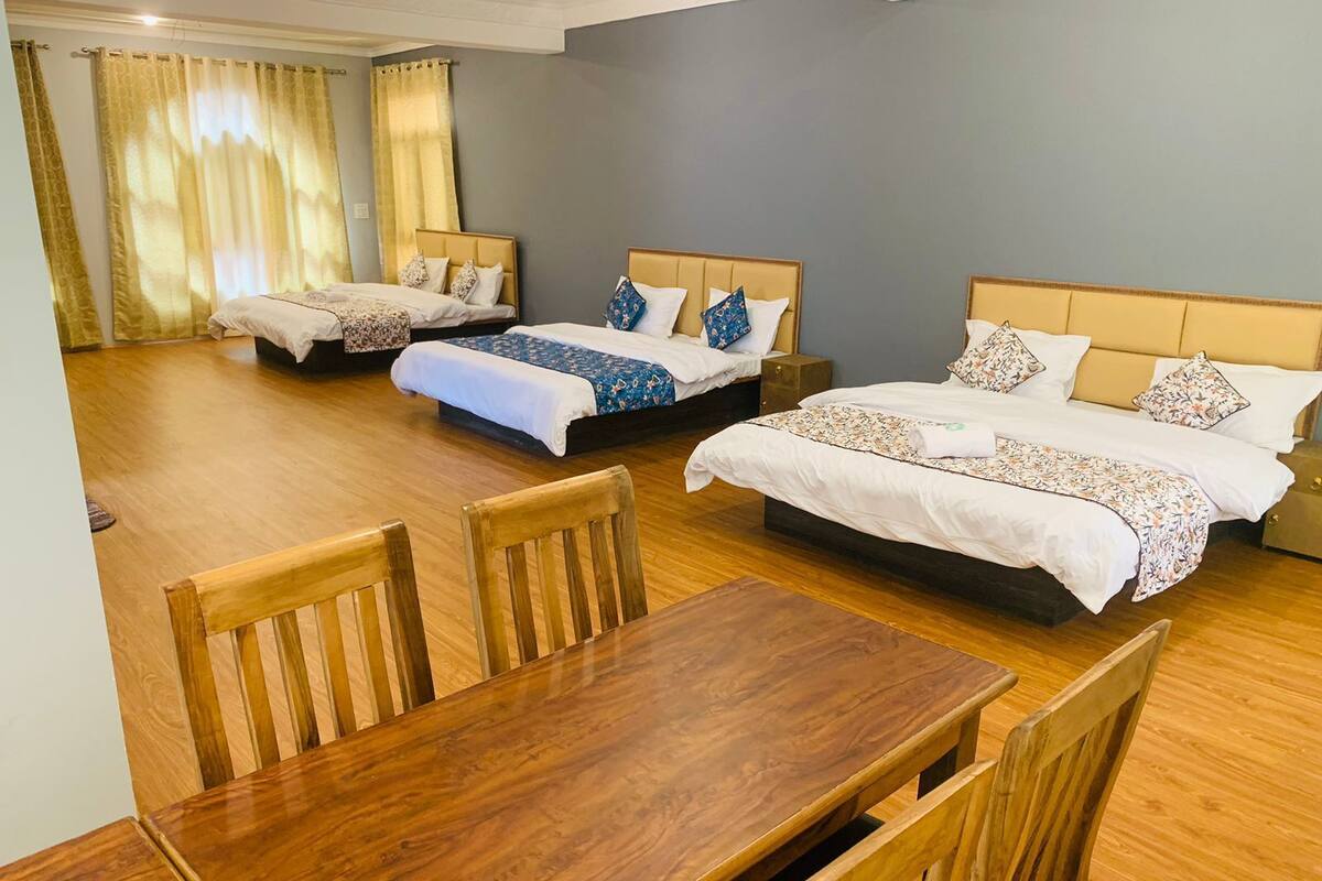 A hotel room with three beds, a wooden dining table, and a large window with curtains.