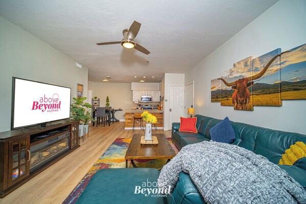 A modern living room with a television, a ceiling fan, a sofa, and a kitchen in the background. - Color Pop by the Pool (Lago Vista)