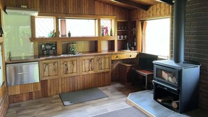 Interior de una cabaña de madera con cocina, comedor y estufa de leña.