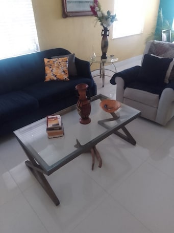 A living room with a glass coffee table, a dark blue sofa, a white armchair, and a vase with flowers on a side table.