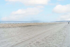 Uma extensa praia de areia com marcas de pneus, céu claro e uma linha de costa distante.