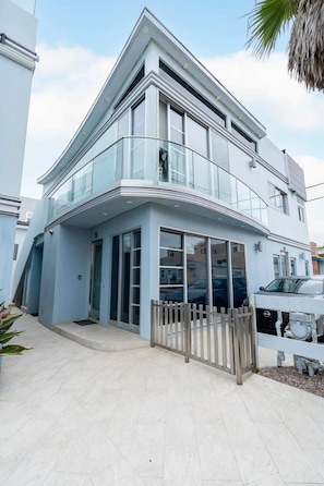 A modern two-story building with a glass balcony, a gated entrance, and a paved driveway.