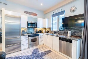 A modern kitchen with stainless steel appliances, white cabinetry, a granite countertop, and a large flat-screen TV.