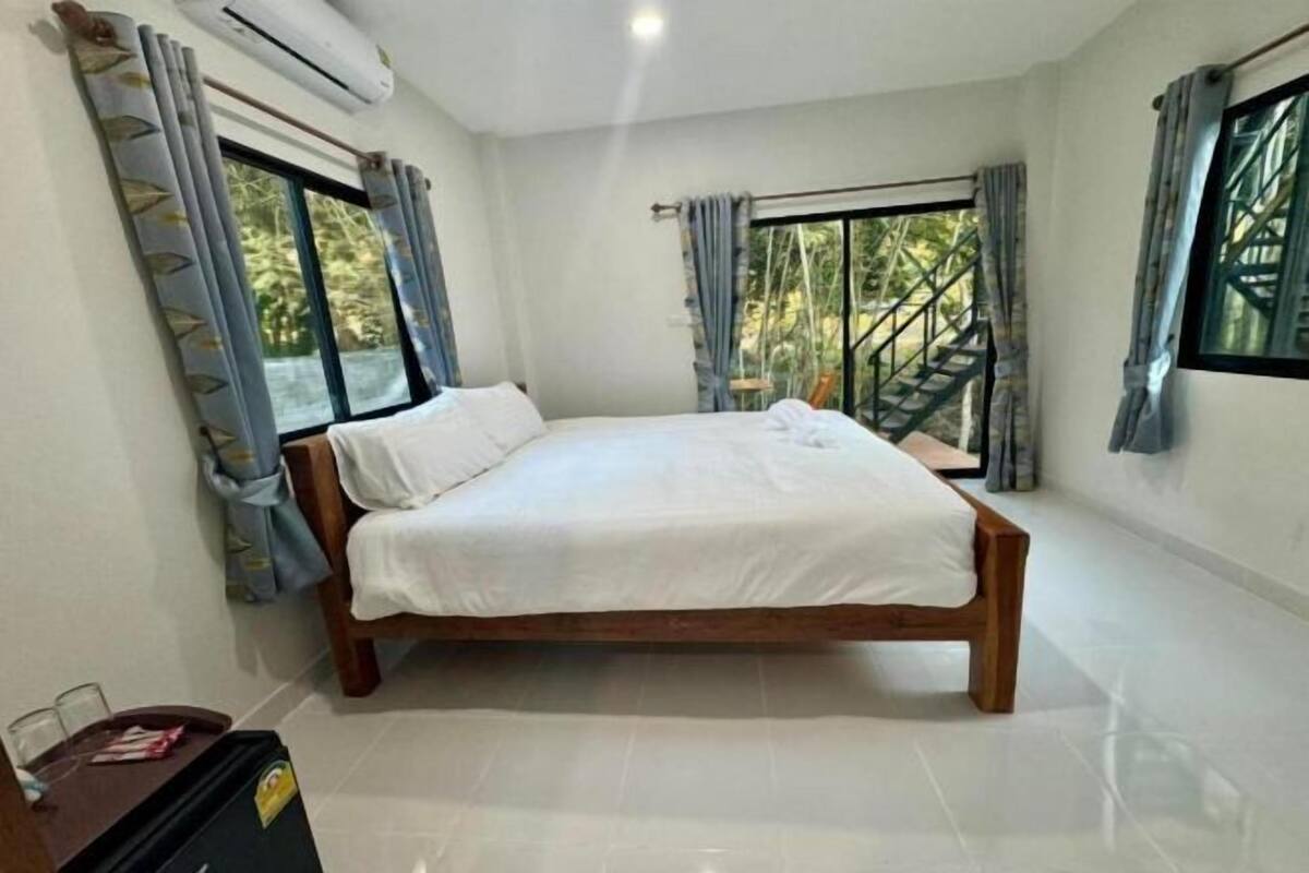A bedroom with a bed, two windows with curtains, an air conditioning unit, and a staircase visible through the window.