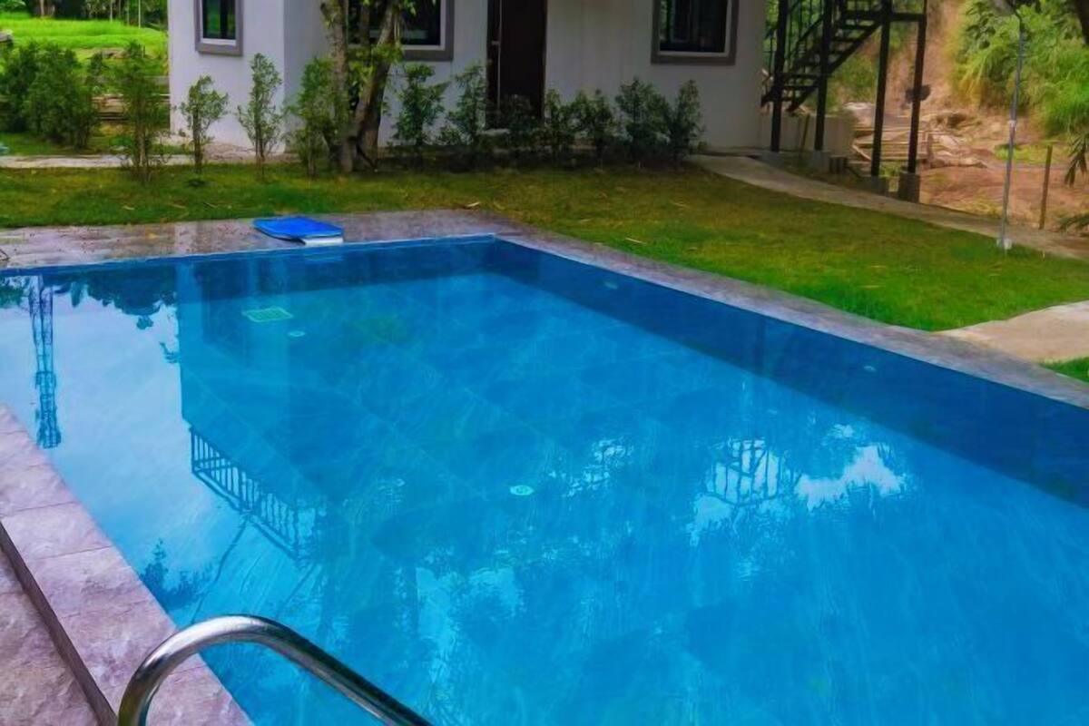 A rectangular swimming pool with a metal ladder, surrounded by a well-maintained lawn and a white building with a black staircase.
