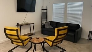 Ein Wohnzimmer mit an der Wand montiertem Fernseher, einer Couch, zwei gelben Sesseln, einem kleinen Tisch und einem Deckenventilator.